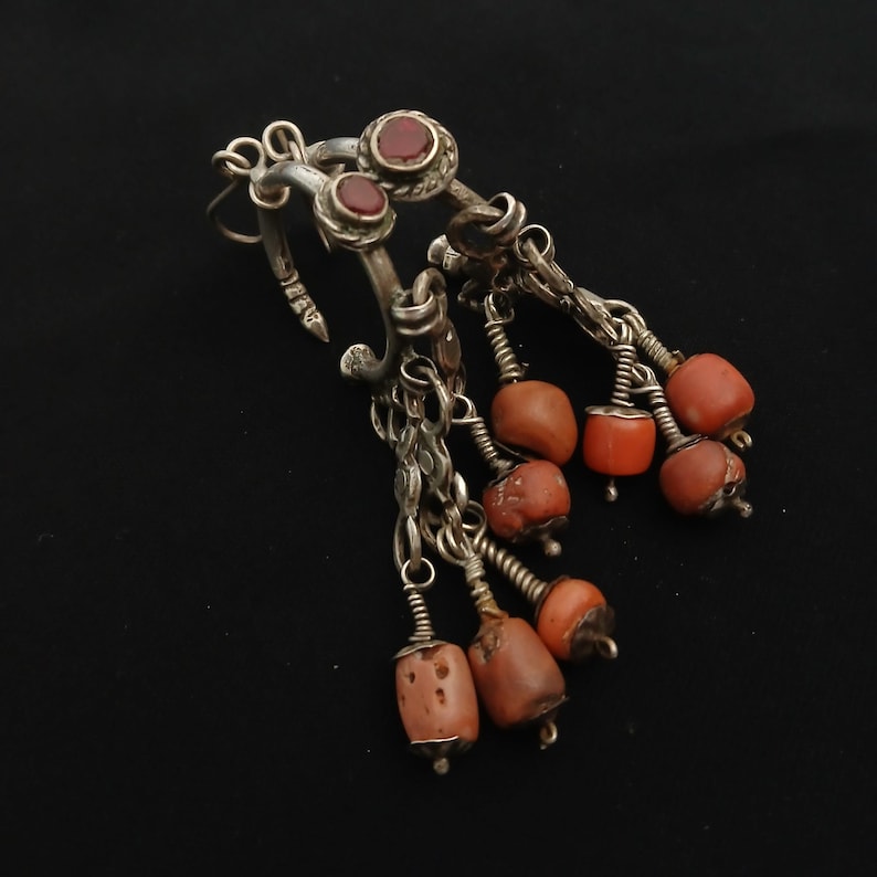 May include: Silver-toned earrings with dangling coral beads. The earrings feature a loop design with three red stone accents. The coral beads are a mix of orange and brown, suspended from delicate chains.