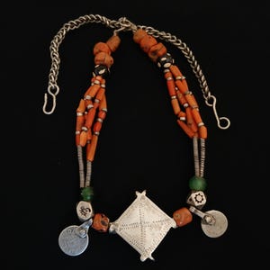 May include: A necklace featuring orange coral beads, silver-toned metal accents, and a central diamond-shaped pendant. The necklace includes small round silver charms and a chain closure. The beads are strung on multiple strands.