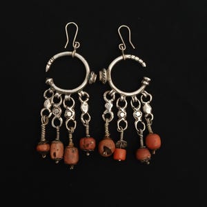 May include: A pair of silver earrings with a crescent-shaped design. Each earring features a series of dangling chains with small, orange-red beads and decorative metal accents. The earrings have a hook closure and are set against a black background.