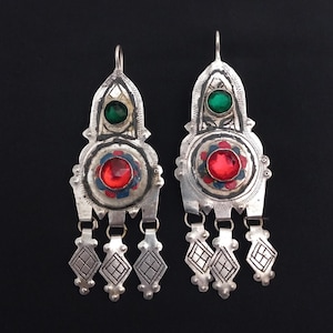 May include: A pair of silver earrings with a green and red gemstone design. Each earring features a green stone at the top, a red stone in the centre, and dangling diamond-shaped charms. The earrings have an antique or vintage aesthetic.