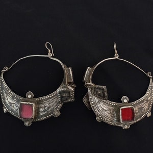 May include: A pair of silver hoop earrings with intricate designs and red glass accents. The earrings are circular and have a crescent moon shape with a decorative pattern.