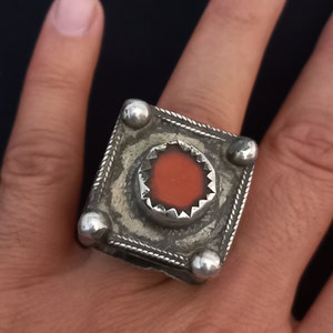Può includere: Un anello d'argento con una montatura quadrata e una pietra rossa al centro. L'anello è decorato con dettagli intricati e ha una finitura testurizzata.