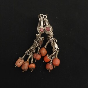 May include: Silver-toned earrings with a dangling design. Each earring features a small, round, red stone at the top, with multiple strands of small chains and orange-red beads hanging below. The earrings are set against a black background.