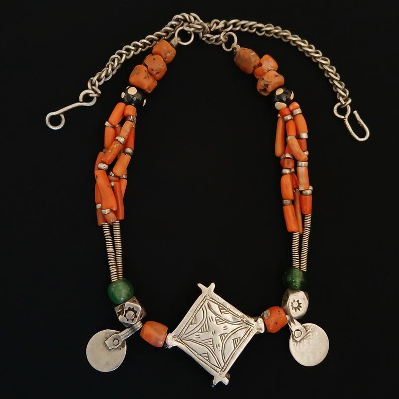 May include: A handcrafted necklace featuring orange coral beads, silver chain, and decorative silver pendants. The necklace has a central diamond-shaped pendant and two round silver pendants. The necklace is displayed against a black background.