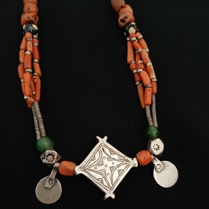 May include: A multi-strand necklace featuring orange coral beads, silver accents, and a central diamond-shaped pendant. The necklace includes small silver disc charms and green glass beads, creating a striking contrast against the black background.