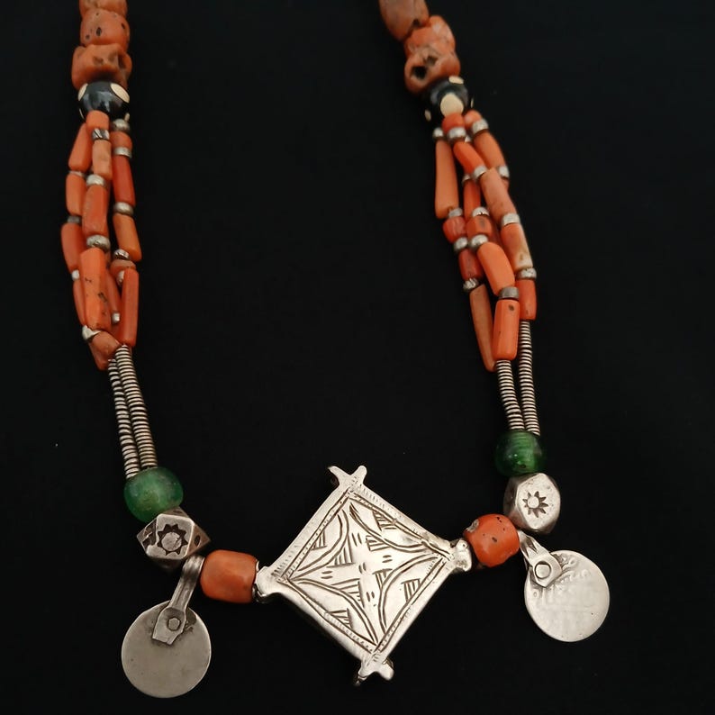 May include: A multi-strand necklace featuring orange coral beads, silver accents, and a central diamond-shaped pendant. The necklace includes small silver disc charms and green glass beads, creating a striking contrast against the black background.