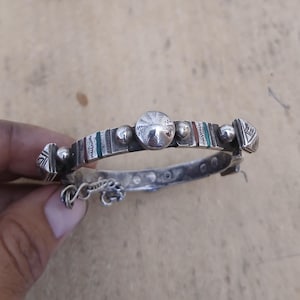 May include: A silver-toned bracelet with intricate detailing. The bracelet features a central sphere and smaller spheres, with sections of red, green, and white stripes. Geometric shapes adorn the ends. A small chain with a decorative element hangs from the bracelet.