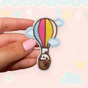Può includere: Toppa ricamata con un design a mongolfiera colorata. Il pallone è diviso in sezioni blu, rosa e gialle. Nuvole bianche circondano il pallone e un cestino marrone è appeso sotto.