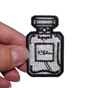 May include: An iron-on patch embroidered in silver and black, depicting a perfume bottle with the word "Chanel" on the label.