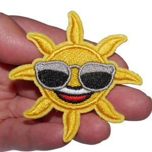 Peut inclure: Un soleil brodé jaune avec un visage souriant portant des lunettes de soleil. Le soleil a une bouche rouge et des dents blanches.