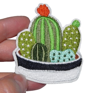 Peut inclure: Un patch brodé blanc représentant une plante de cactus en pot avec des détails verts, orange et bruns. Le cactus est dans un pot blanc avec un bord noir.