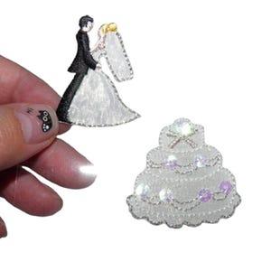 Peut inclure: Deux patchs thermocollants brodés blancs, l'un représentant un marié et une mariée, l'autre un gâteau de mariage blanc avec des paillettes irisées.