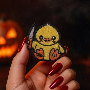 Könnte beinhalten: Gestickter Aufnäher mit einer gelben Cartoon-Ente, die ein Messer hält. Der Aufnäher hat schwarze Umrisse und die Worte "Duck You" sind auf die Füße der Ente gestickt. Der Hintergrund ist verschwommen, mit einem Halloween-Thema.