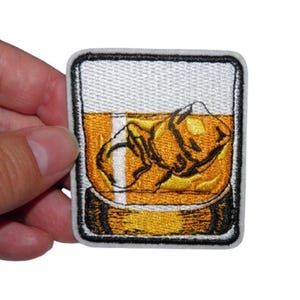 Peut inclure: Écusson brodé représentant un verre de whisky carré avec des glaçons. Le verre est rempli de liquide ambré et a une bordure noire et blanche. L'écusson est tenu par une main.