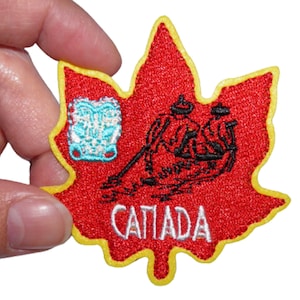 Könnte beinhalten: Roter Ahornblatt-förmiger Patch mit einem Schwarzweißbild eines Kanus mit zwei paddelnden Personen. Das Wort "CANADA" ist in Weiß auf den Patch gestickt. Der Patch hat einen gelben Rand.