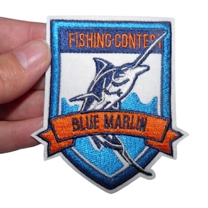 May include: A white embroidered patch featuring a blue marlin fish leaping from the water. The patch has a blue and white shield with the text "FISHING CONTEST" at the top and "BLUE MARLIN" on an orange ribbon at the bottom.