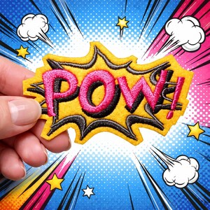Peut inclure: Un écusson "POW!" jaune, noir et rose de style bande dessinée. L'écusson présente un fond jaune avec des lignes noires en zigzag et du texte brodé en rose. L'arrière-plan est un design de bande dessinée coloré avec des étoiles et des bulles.