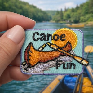 Puede incluir: Parche bordado con las palabras "Canoe Fun" y un diseño de canoa. El parche presenta un fondo azul claro, un sol amarillo y una canoa en tonos naranja y marrón. El parche se sostiene en una mano.