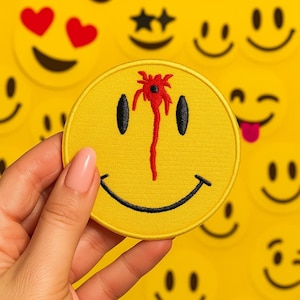 May include: A yellow smiley face patch with black eyes and a curved mouth, featuring a red blood-like splatter design. The patch is round and surrounded by other yellow smiley face patches with various expressions.