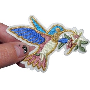 Peut inclure: Patch brodée thermocollante représentant un colibri aux accents bleus, roses et dorés, perché sur une branche avec une fleur blanche.