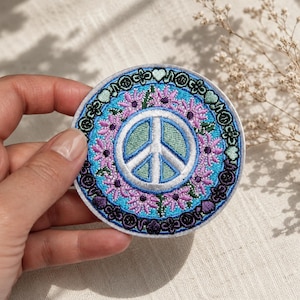 May include: A circular embroidered patch featuring a central white peace sign on a light green background. The design is surrounded by a ring of pink daisy-like flowers on a bright blue field, finished with a dark purple border of small hearts.