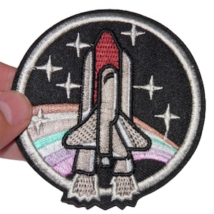 May include: A black and white embroidered patch of a space shuttle with a rainbow stripe and stars. The patch is circular and has a white border.