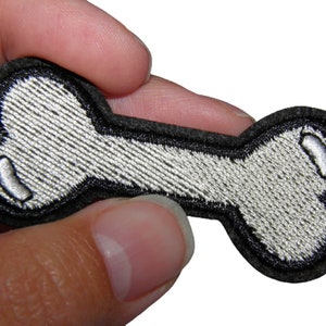 Peut inclure: Un patch en forme d'os blanc avec des coutures noires autour des bords. Le patch est en feutre et a une surface texturée.