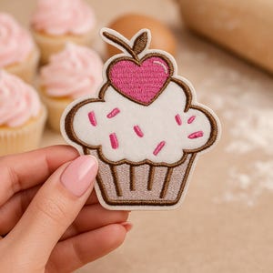 Könnte beinhalten: Gestickter Cupcake-Aufnäher mit weißem Zuckerguss, rosa Streuseln und einem rosa Herz. Der Aufnäher hat eine braune Umrandung und wird in einer Hand gehalten. Cupcakes mit rosa Zuckerguss sind im Hintergrund.
