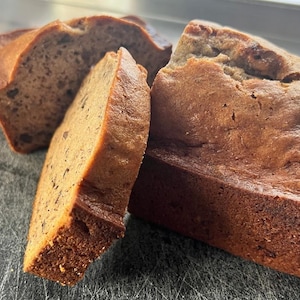 Könnte beinhalten: Drei Scheiben Bananenbrot auf einem grauen Schneidebrett. Das Brot ist goldbraun und hat eine leicht krümelige Textur.