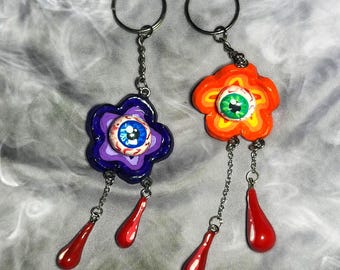 May include: Two keychains with colorful flower charms. Each flower has a large eye painted on it. One flower is orange, yellow, and green. The other flower is purple, blue, and red. Both keychains have red dangling charms.