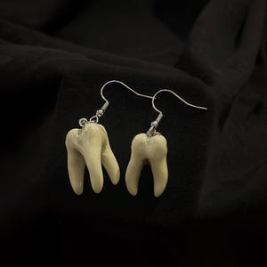 Pode incluir: Um par de brincos de prata com pingentes em forma de dentes brancos.