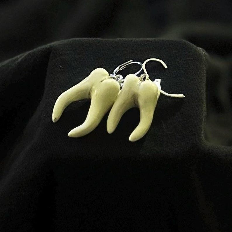 Pode incluir: Um par de brincos com tr&ecirc;s pingentes brancos em forma de dente pendurados em ganchos prateados.