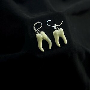 Pode incluir: Um par de brincos de prata com pingentes de dentes brancos. Os dentes t&ecirc;m um design realista e est&atilde;o pendurados nos brincos.