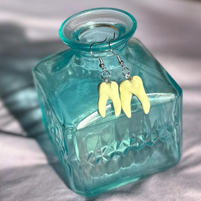 Pode incluir: Um par de brincos amarelos em forma de dente pendurados em ganchos prateados. Os brincos s&atilde;o exibidos em uma garrafa de vidro azul.