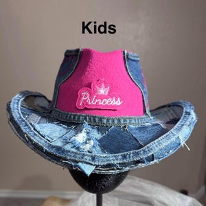 Può includere: Un cappello da cowboy in denim con una corona rosa e la parola "Princess" ricamata sul davanti. Il cappello è realizzato in denim patchwork e ha una corona rosa. La parola "Kids" è stampata in alto.