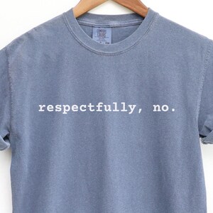 May include: A dusty blue t-shirt with the phrase "respectfully, no." printed in white. The short-sleeved shirt is hanging on a wooden hanger against a white background. The sleeves are cuffed.