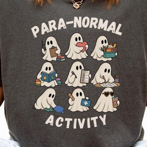 Peut inclure: Un t-shirt gris anthracite à manches courtes avec une impression graphique de neuf fantômes blancs effectuant diverses activités. Le texte "PARA-NORMAL ACTIVITY" est imprimé en blanc au-dessus et au-dessous des fantômes, représentés en train de lire, de tenir des objets et de porter des lunettes de soleil.