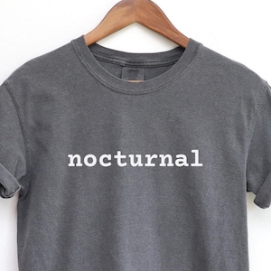 May include: Dark gray t-shirt with the word "nocturnal" printed in white. The shirt has rolled-up sleeves and is hanging on a wooden hanger against a white background. The t-shirt is a casual wear item.