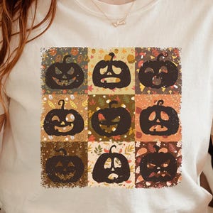 May include: A cream-colored short-sleeve t-shirt featuring a square graphic print. The design consists of nine individual panels, each displaying a dark brown silhouette of a jack-o'-lantern with a unique facial expression against autumn-themed backgrounds.