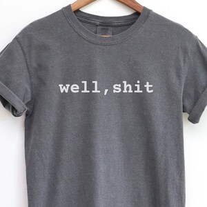 May include: Dark gray t-shirt with the phrase "well, shit" printed in white. The shirt has rolled-up sleeves and a crew neck. The garment is hanging on a wooden hanger against a white background.