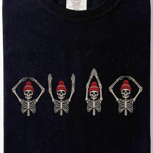 May include: A black crew neck t-shirt featuring a graphic print of four skeletons wearing red beanies. Each skeleton is positioned in a different pose, with their arms forming the letters C, O, D, and E. The shirt has a Comfort Colors brand label.