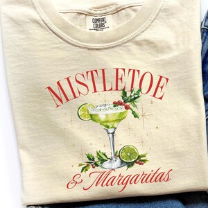 Pu&ograve; includere: Una t-shirt color crema con la grafica di un margarita verde in un bicchiere con una fetta di lime, bacche rosse e foglie verdi. Il testo MISTLETOE & MARGARITAS &egrave; stampato in rosso sopra e sotto l'illustrazione del cocktail.