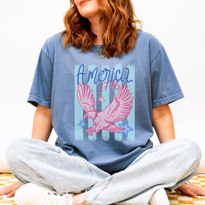 May include: A blue t-shirt featuring a pink eagle graphic with the word "America" and the year "1776". The design includes blue stripes and stars. The shirt is a casual style.
