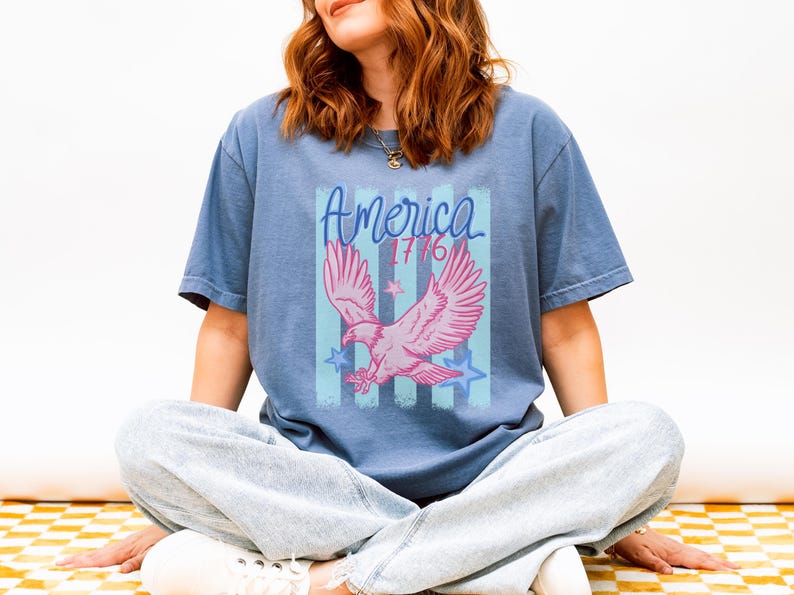 May include: A blue t-shirt featuring a pink eagle graphic with the word "America" and the year "1776". The design includes blue stripes and stars. The shirt is a casual style.
