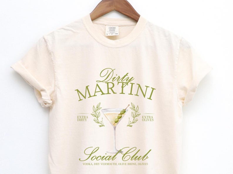 May include: A cream-coloured t-shirt featuring a green graphic of a martini glass with olives. The text reads Dirty Martini Social Club, Extra Dirty, Extra Olives, and Vodka, Dry Vermouth, Olive Brine, Olives. The shirt has short sleeves and a crew neckline.