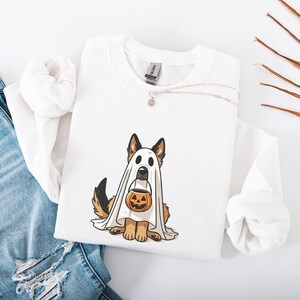 May include: White sweatshirt featuring a German Shepherd dog dressed as a ghost, holding a jack-o'-lantern. The dog has brown and black fur. A silver necklace with a small pendant is on the sweatshirt. Blue jeans are in the background.