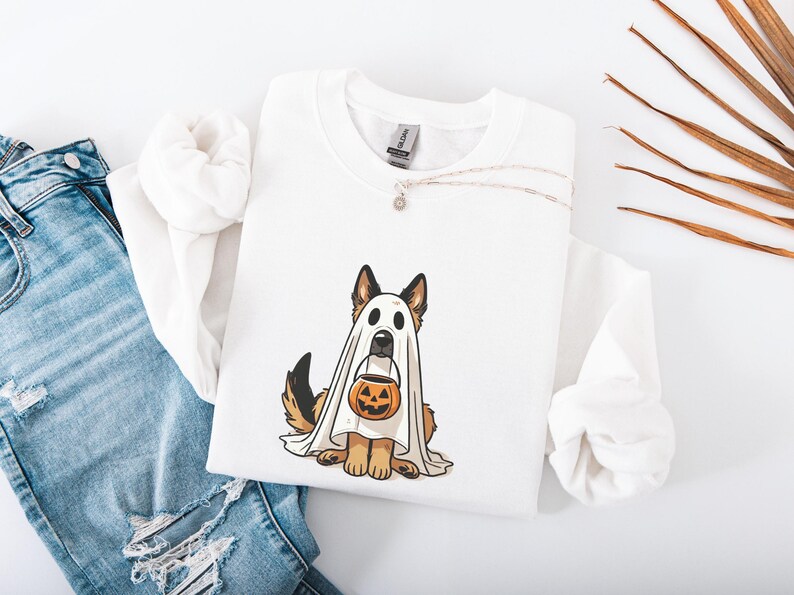 May include: White sweatshirt featuring a German Shepherd dog dressed as a ghost, holding a jack-o'-lantern. The dog has brown and black fur. A silver necklace with a small pendant is on the sweatshirt. Blue jeans are in the background.