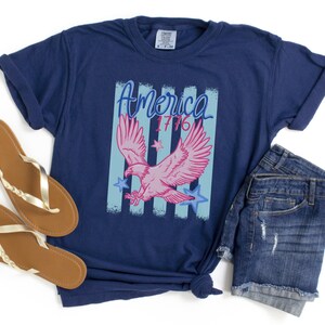 May include: Navy blue t-shirt with a graphic featuring a pink eagle, blue stars, and the text "America 1776". The shirt is paired with denim shorts and brown and gold sandals.