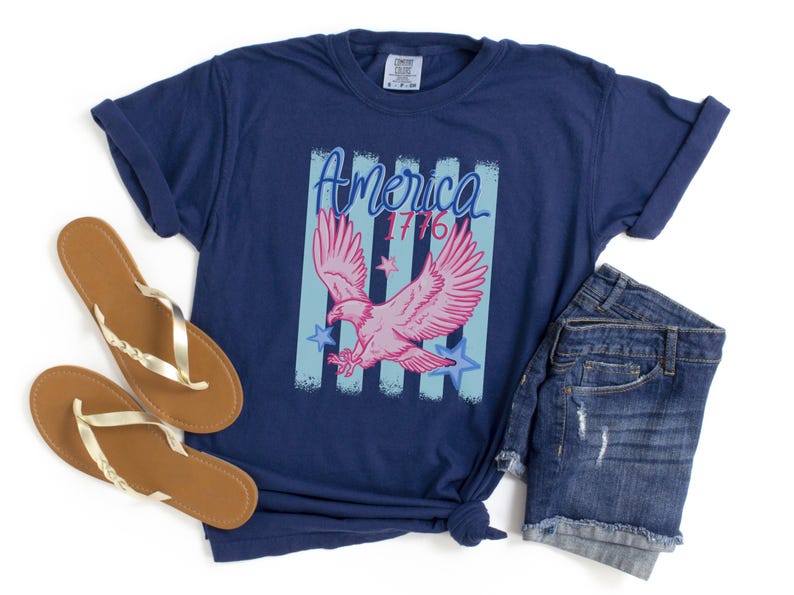 May include: Navy blue t-shirt with a graphic featuring a pink eagle, blue stars, and the text "America 1776". The shirt is paired with denim shorts and brown and gold sandals.