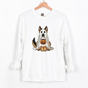 May include: White sweatshirt featuring a cartoon dog dressed as a ghost for Halloween. The dog is wearing a white sheet with eye holes and holding a jack-o'-lantern bucket. The dog has brown and black fur and is sitting down.